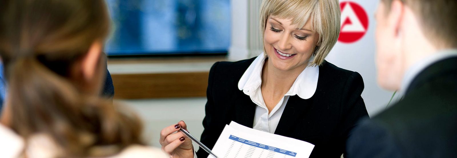 Auf dem Bild ist eine Frau im Anzug zu sehen, die mit einem Stift auf eine Tabelle auf einem Blatt Papier zeigt.