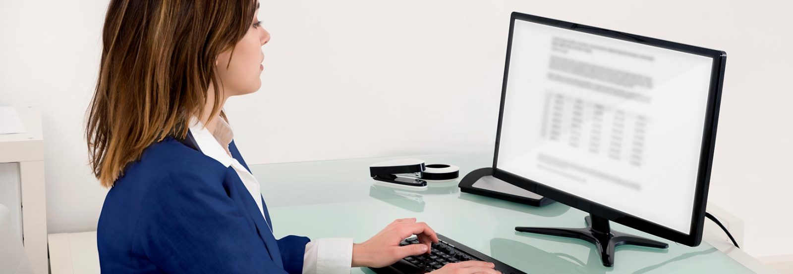 The picture shows a woman dressed in a blazer at a desk in front of a computer.