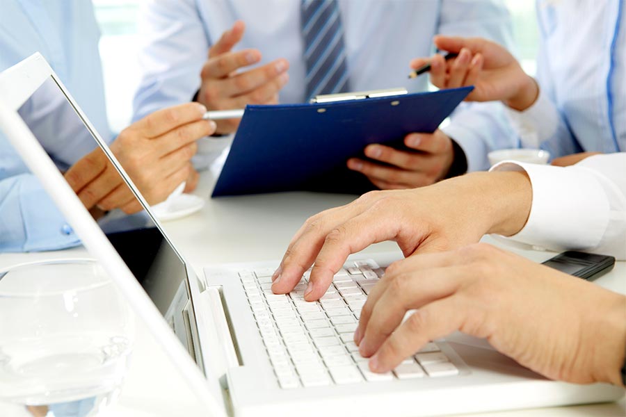 Das Bild zeigt Hände einer Person, wie sie auf einer Laptop-Tastatur tippen und im Hintergrund die Hände dreier Personen, die über Aufzeichnungen diskutieren