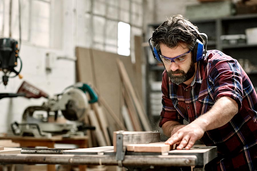Das Bild zeigt einen Tischler im karierten Hemd, mit Schutzbrille und Ohrenschützern, der ein Holzbrett bearbeitet.