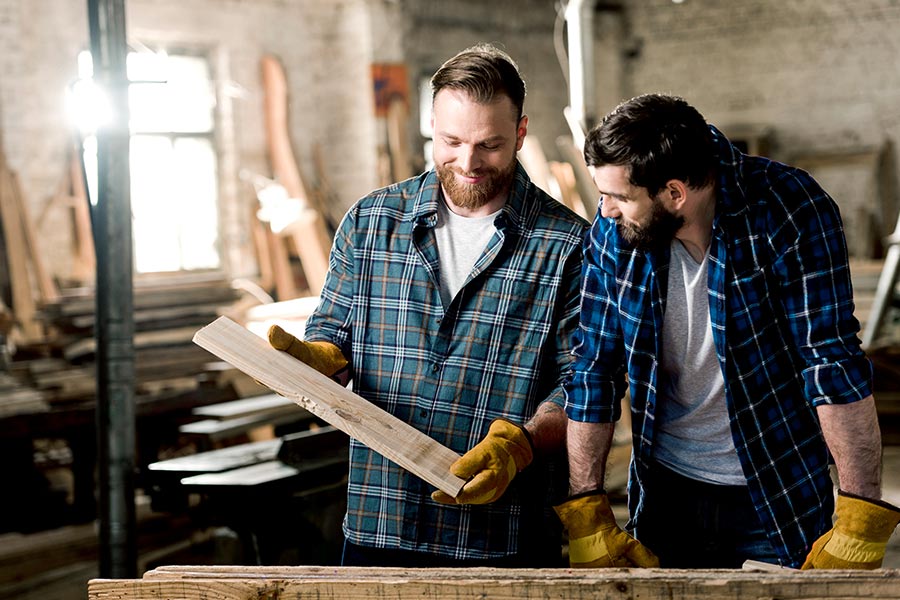 Das Bild zeigt zwei Männer in karierten Hemden in einer Werkstatt, wobei der eine ein Holzbrett in den Händen hält.