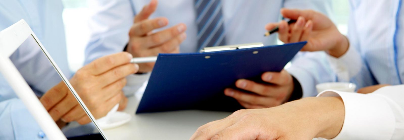 Das Bild zeigt Hände einer Person, wie sie auf einer Laptop-Tastatur tippen und im Hintergrund die Hände dreier Personen, die über Aufzeichnungen diskutieren