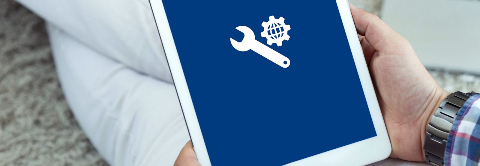 Das Bild zeigt eine auf dem Boden sitzende Person, die auf dem Schoß ein Tablet hält auf dessen Display ein Werkzeug-Icon abgebildet ist.