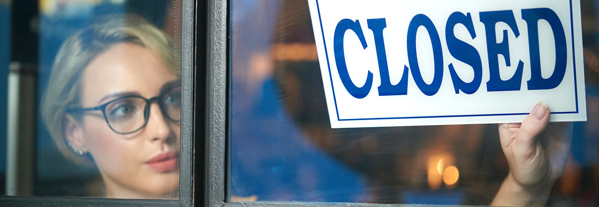 The picture shows a woman wearing glasses, who is turning the “Closed” sign at the door over.