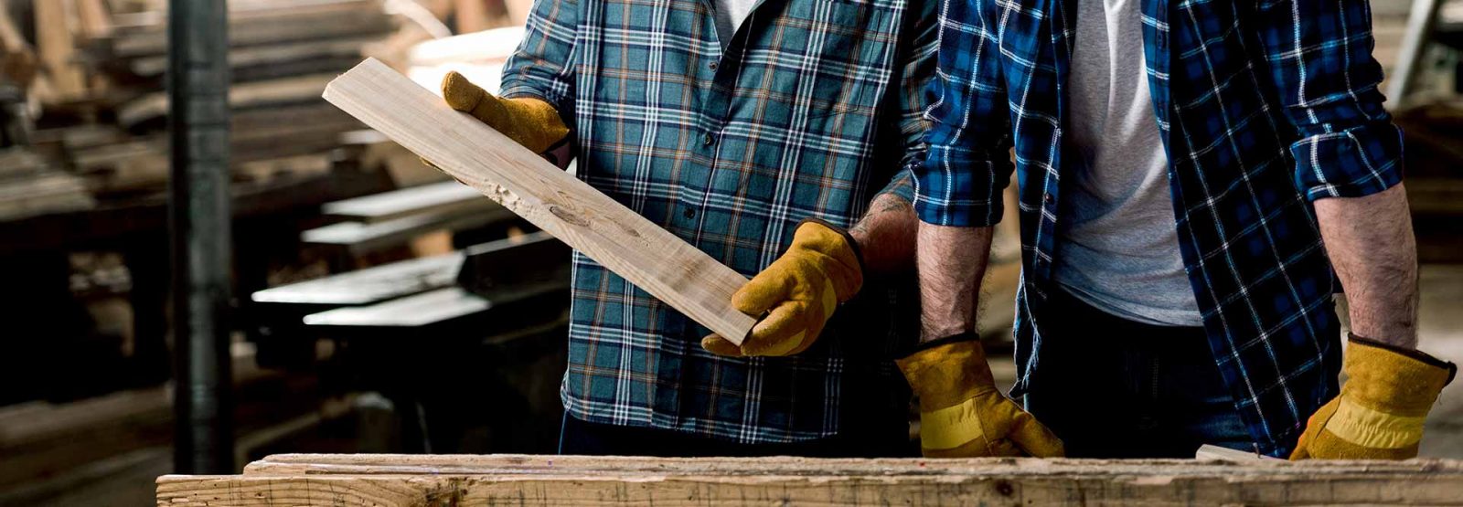 The picture shows two men wearing lumberjack shirts in a workshop; one of them is holding a wooden board in his hands.