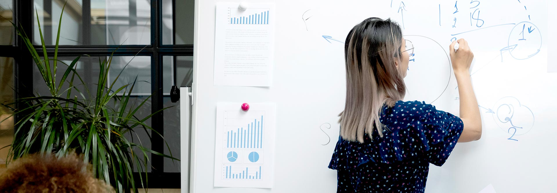 The picture shows a woman in a meeting, who is writing on a pin board with diagram printouts attached to it.