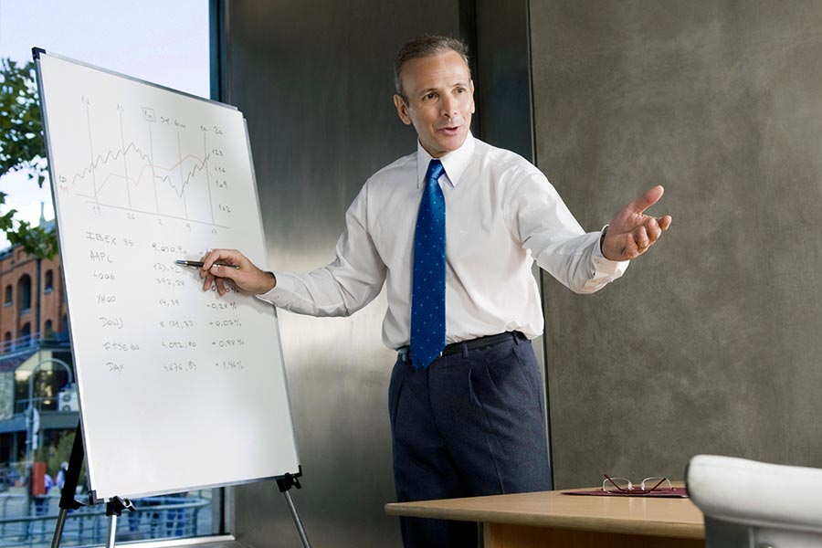 The picture shows a man wearing a tie and formal trousers, who is standing in front of a flip chart and explaining his notes on it.