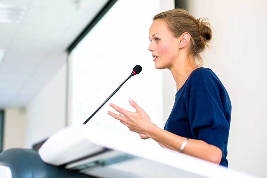 The picture shows a woman behind a speaker’s desk holding a lecture via the microphone.